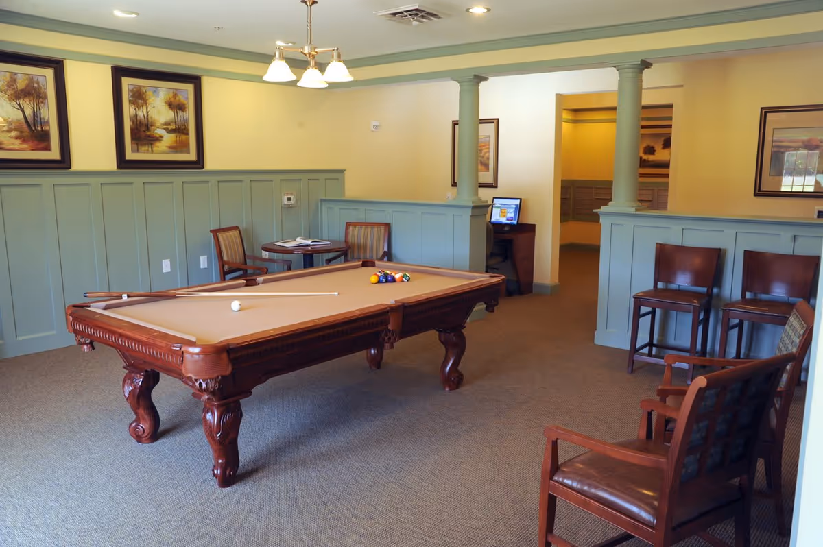 A well-lit room with a pool table in the center, surrounded by wooden chairs. The walls are painted light green with wainscoting, and there are framed landscape paintings hanging on the walls. A small round table with two chairs is in the corner, and a computer workstation is visible through an open doorway.