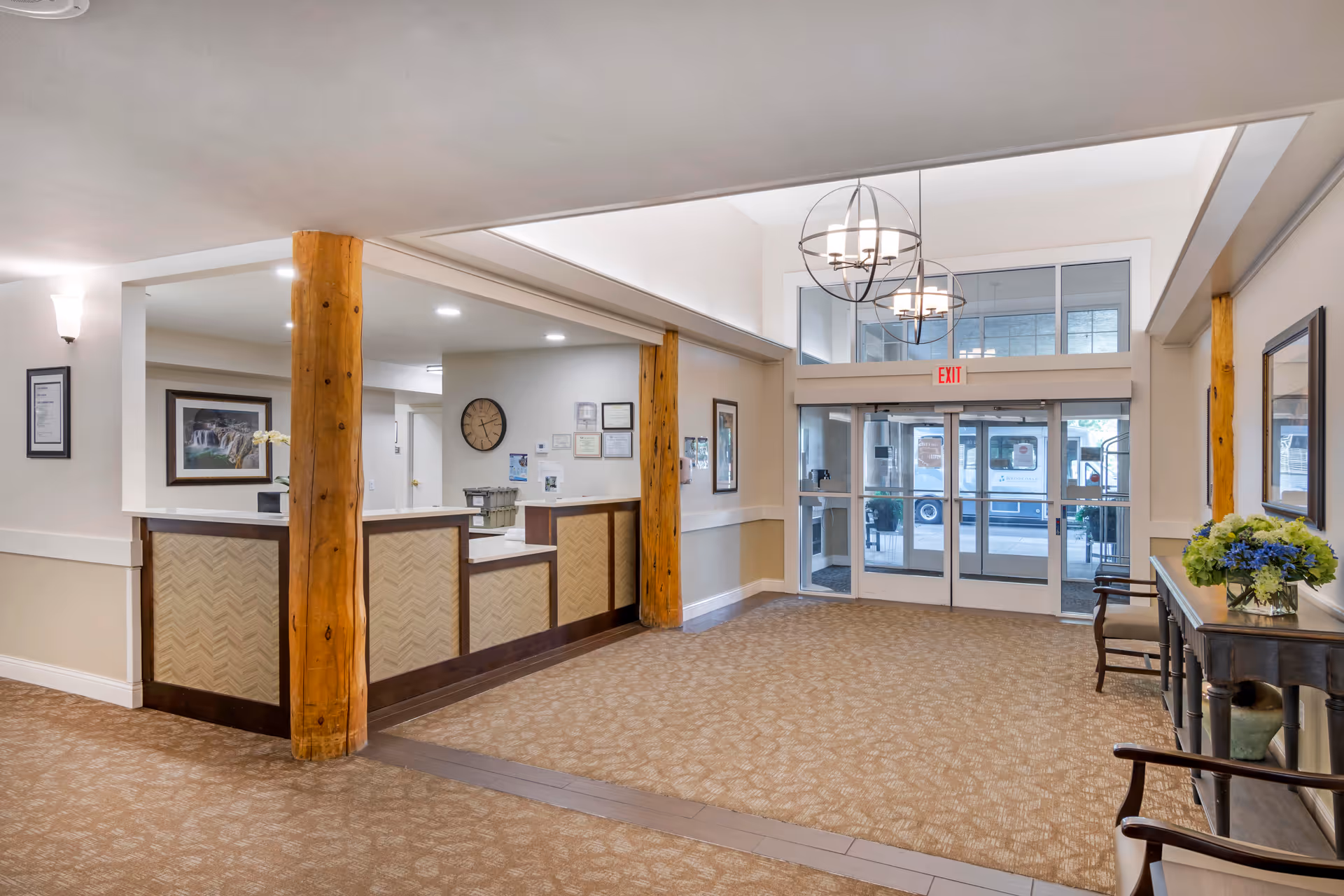 Interior view of a senior living facility lobby with a reception desk supported by wooden pillars, beige patterned carpet, a table with a flower arrangement, chairs, and glass double doors leading outside. The area is well-lit with modern hanging light fixtures and wall sconces.
