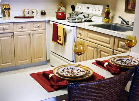 A cozy kitchen with light wood cabinets, a white stove with a red kettle on top, a sink, and a small dining area with two place settings including decorative plates, red napkins, and amber-colored wine glasses.