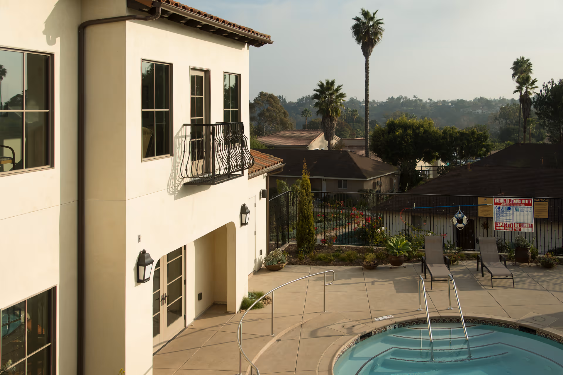 Outdoor view of a senior living facility showing a small pool or hot tub with metal handrails, several lounge chairs, potted plants, and a beige building with multiple windows and a small balcony. In the background, there are palm trees, other buildings, and a fenced garden area.