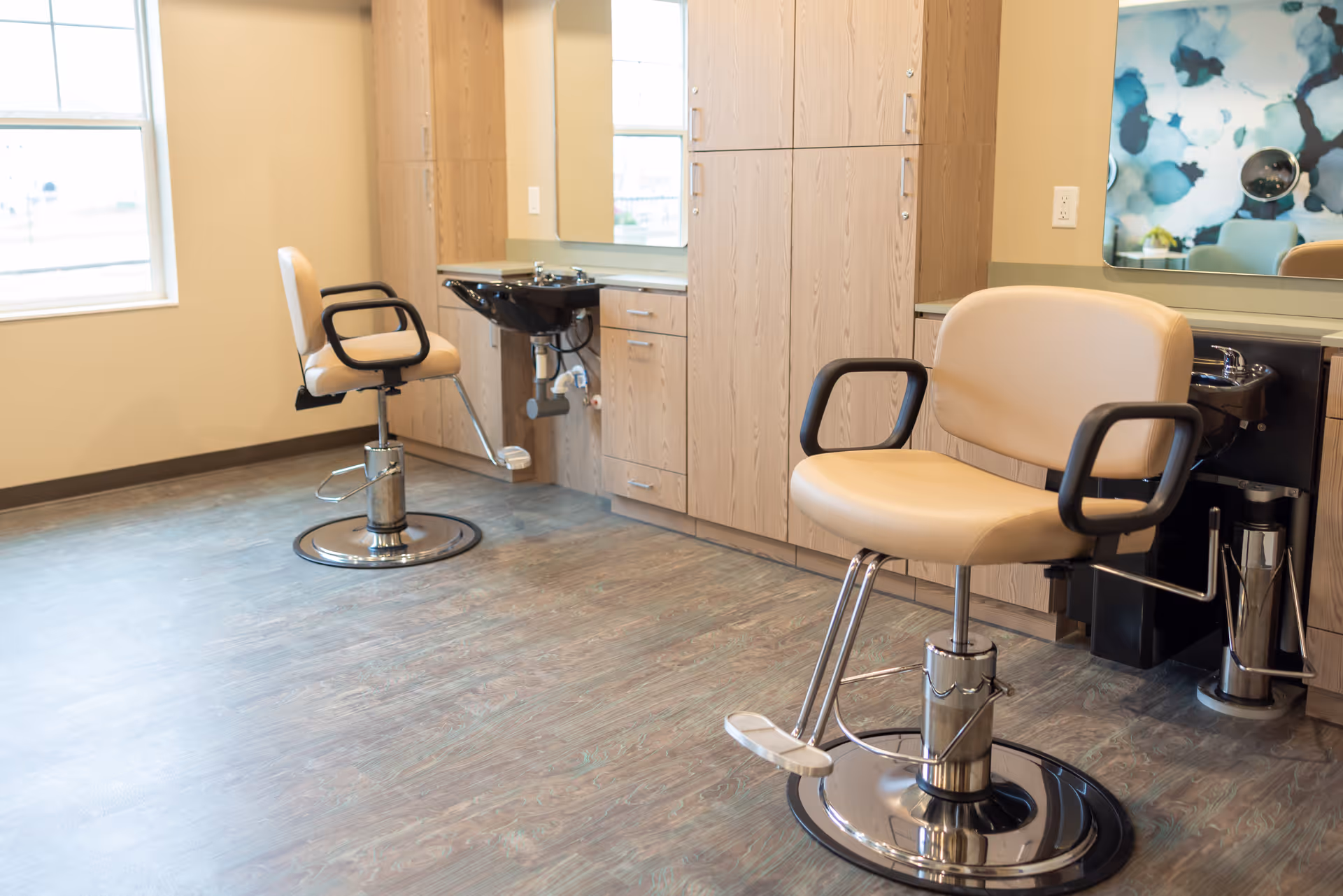 Two salon chairs and hair-washing sinks in a bright salon area with wood cabinets and a large mirror.