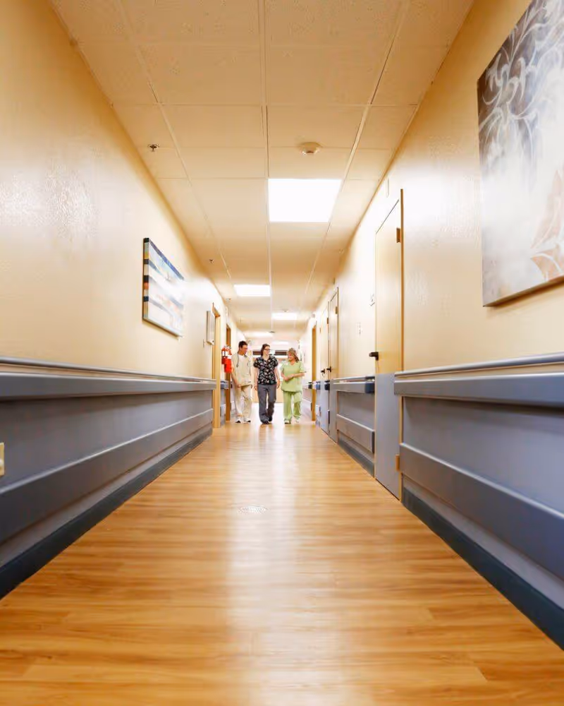 Well-lit senior care hallway with wood-look flooring and three staff walking toward the camera.