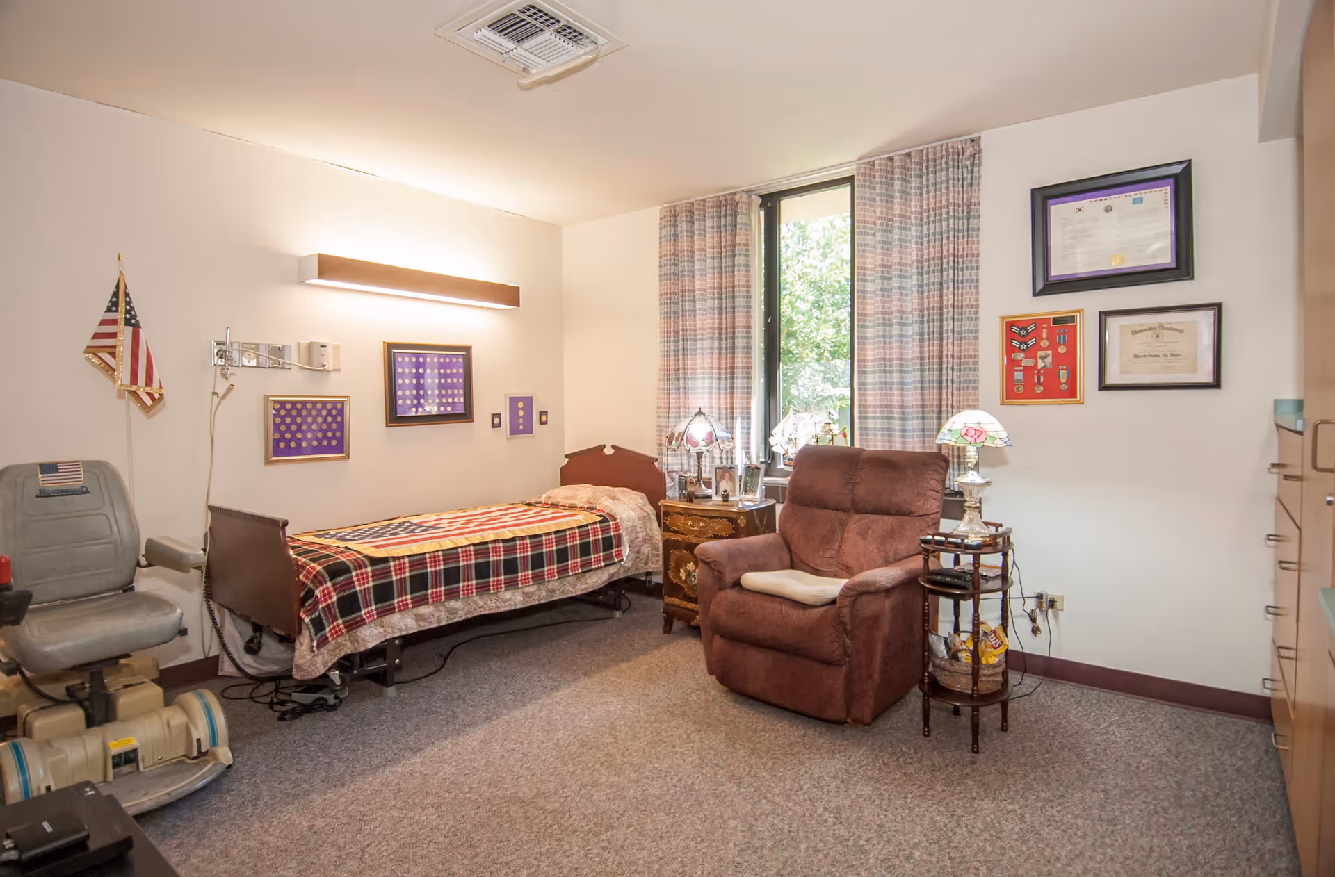 A cozy nursing home bedroom with a single bed covered by a plaid blanket and an American flag blanket. There is a brown recliner chair with a cushion, a small wooden side table with a lamp, and framed certificates and medals on the walls. A window with patterned curtains lets in natural light, and a mobility scooter is parked near the bed.