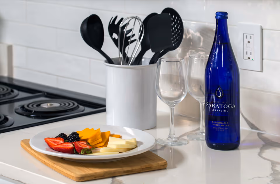 A kitchen countertop with a white plate holding sliced strawberries, blackberries, and two types of sliced cheese on a wooden cutting board. Behind the plate, there is a white utensil holder containing black kitchen utensils, two empty wine glasses, and a blue bottle of Saratoga sparkling carbonated spring water. The background features a white tiled backsplash and an electric stove with coil burners.