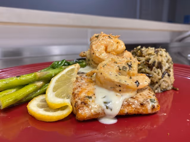 A plated meal featuring grilled salmon topped with creamy shrimp, served with asparagus, lemon slices, and a side of wild rice on a red plate.