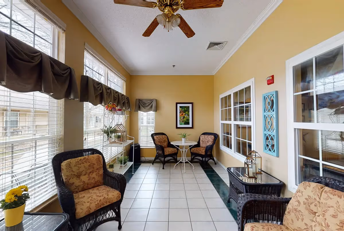 A bright sunroom with large windows on two sides, featuring wicker chairs with floral cushions, small tables with decorative plants and lanterns, a ceiling fan with lights, and beige walls adorned with framed artwork and decorative wall hangings.