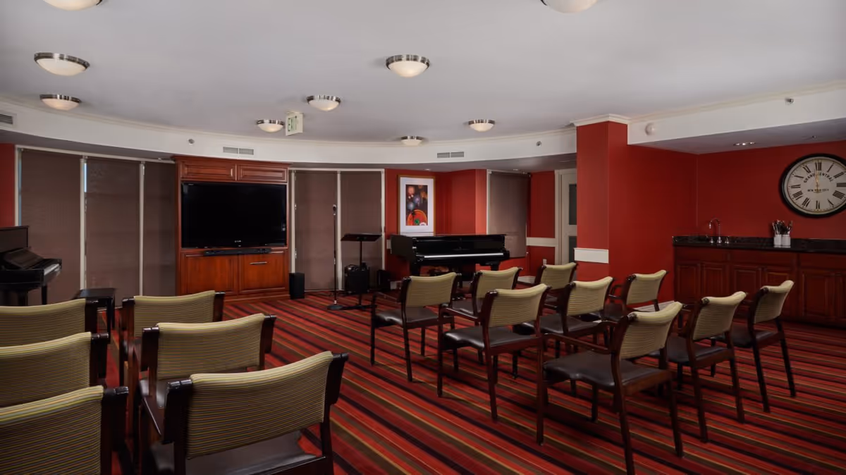 Small auditorium-style common room with rows of chairs facing a television and a piano, red walls and striped carpet.