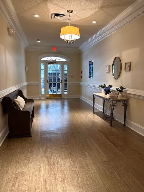 A well-lit hallway in a senior living facility with wooden flooring, beige walls, and white trim. On the left side, there is a wooden bench with a white pillow. On the right side, a narrow table holds a telephone and some decorative items, with a round mirror and framed pictures hanging above it. At the end of the hallway, there is a glass door with a view of a parked vehicle outside.