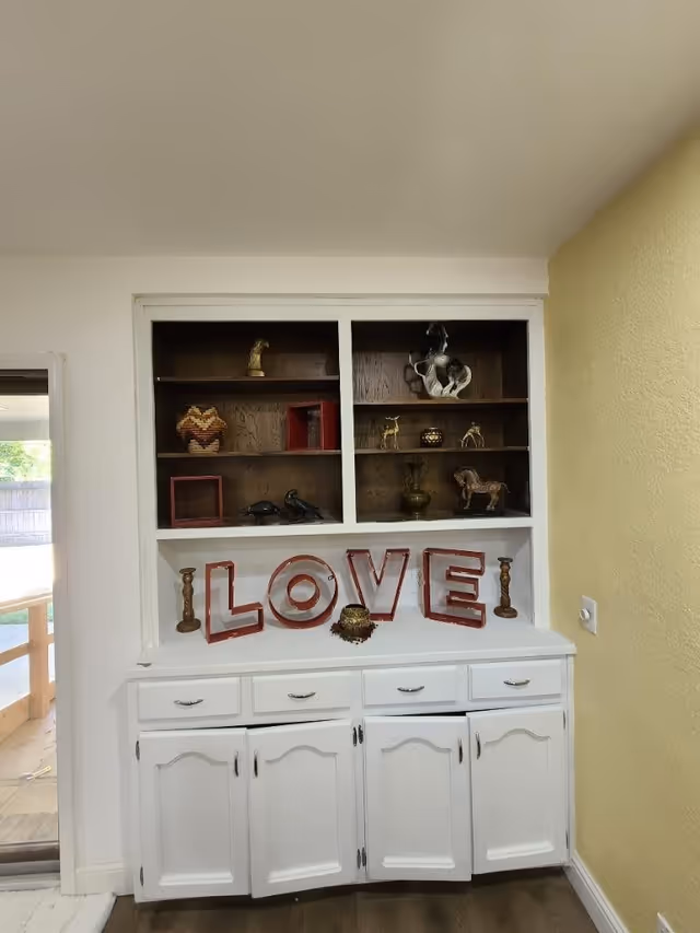 A white built-in cabinet with shelves displaying decorative items including small sculptures and candle holders. Large letters spelling 'LOVE' are placed on the middle shelf. The cabinet is set against a wall with a yellow section on the right and a sliding glass door on the left.