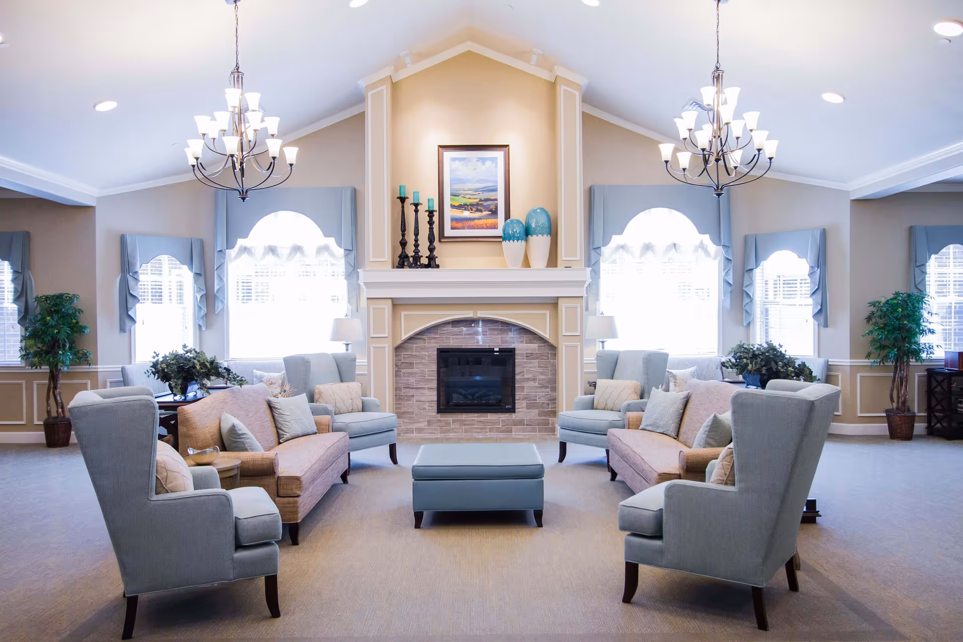 A bright and spacious living room with a high vaulted ceiling, featuring two chandeliers, a central fireplace with decorative vases and candles on the mantel, two beige sofas, four light blue armchairs, a blue ottoman, and large windows with blue valances allowing natural light to fill the room.