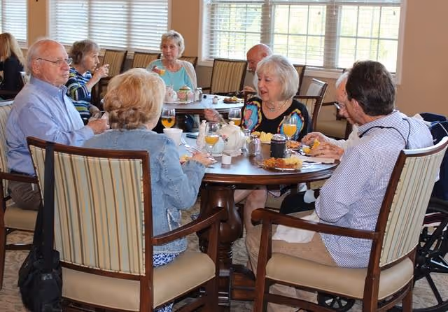 A group of elderly people sitting around a round dining table in a well-lit room with large windows, enjoying a meal and drinks together.