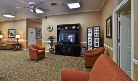 A senior living facility common area with beige walls and carpeted floor. The room features a large black entertainment center with a TV, two orange armchairs, a matching orange sofa, framed pictures on the walls, a potted plant, and ceiling fans with lights.