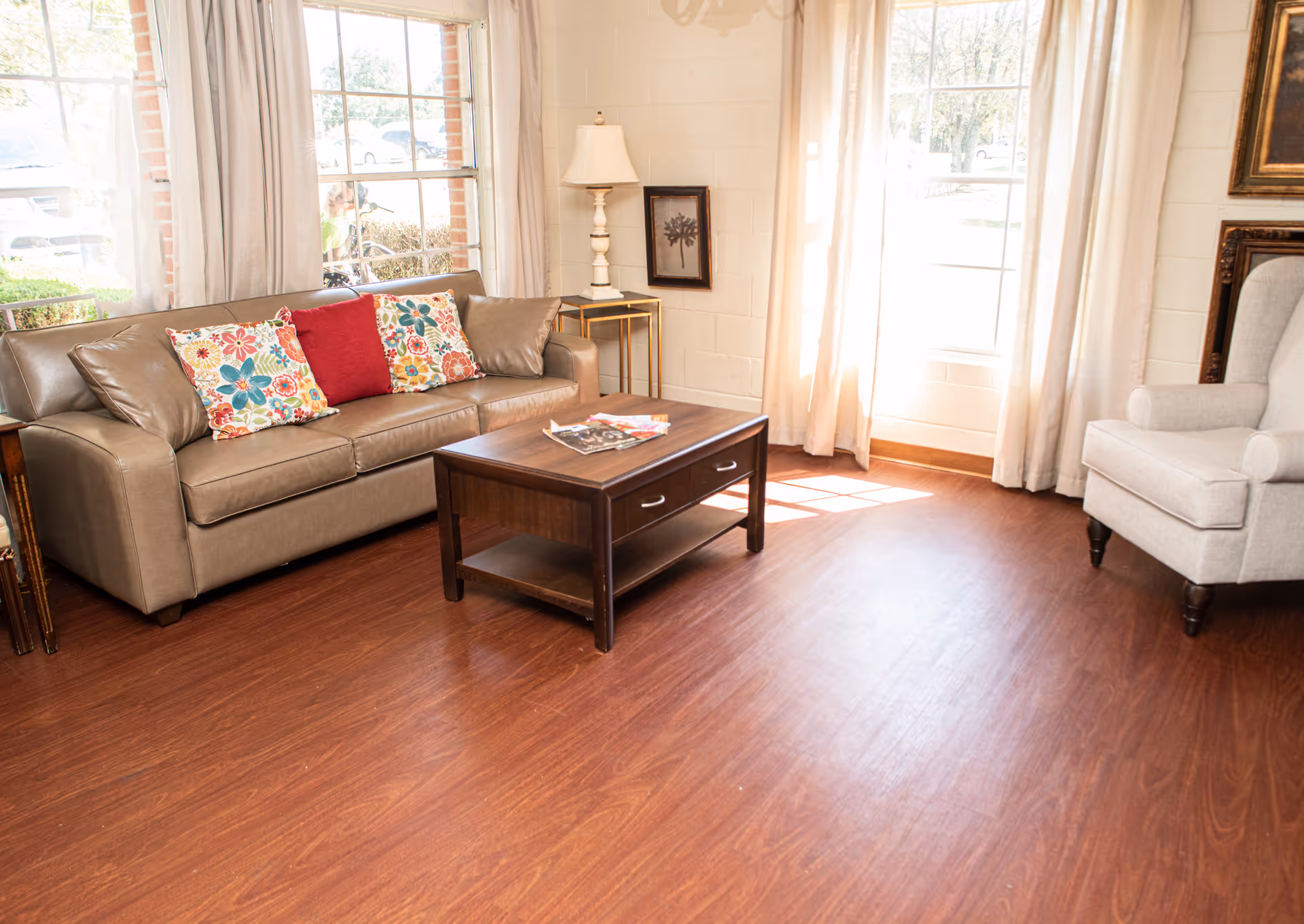 A bright living room with a brown leather sofa adorned with colorful floral and red pillows, a wooden coffee table with magazines on top, a beige armchair, a floor lamp on a small side table, and large windows with white curtains letting in natural light.