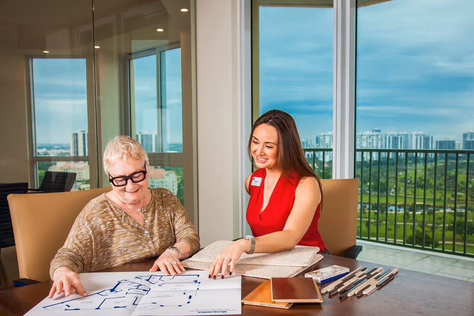 An elderly woman and a younger woman sitting at a table in a bright room with large windows overlooking a cityscape and greenery. They are looking at a floor plan and fabric samples, discussing interior design or housing options. The younger woman is wearing a red sleeveless top and a name badge.