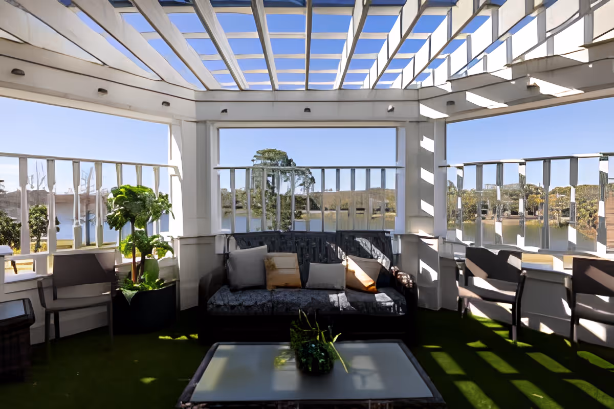 A bright outdoor seating area with a white pergola overhead, featuring a cushioned sofa with pillows, several chairs, a glass-top coffee table with a small plant centerpiece, and a view of trees and water in the background.