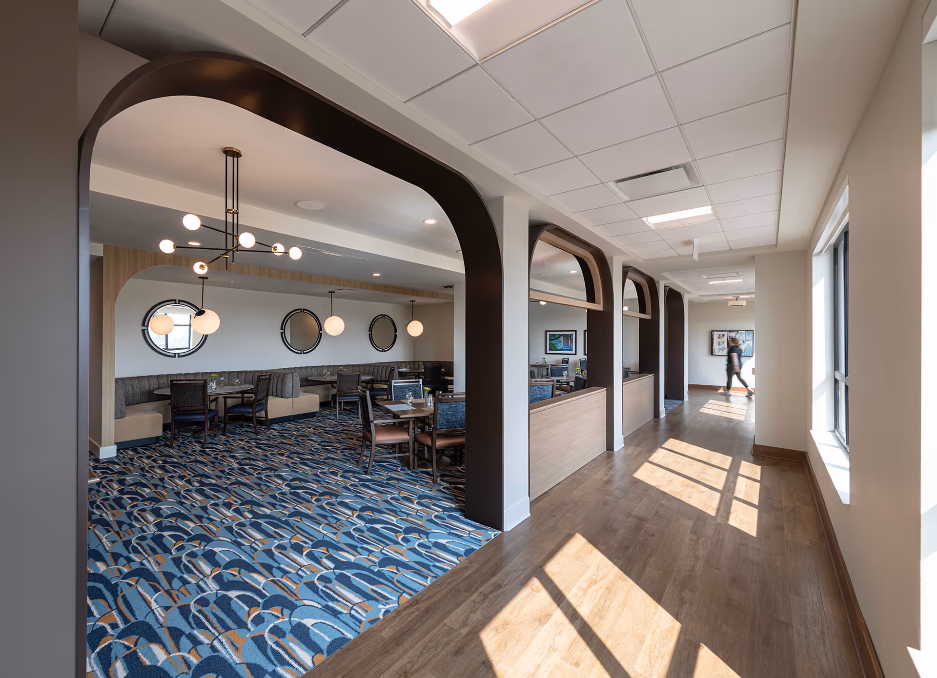 A sunlit corridor opens into a dining/lounge area with arched wood accents, round pendant lights, booths and tables on patterned carpet.