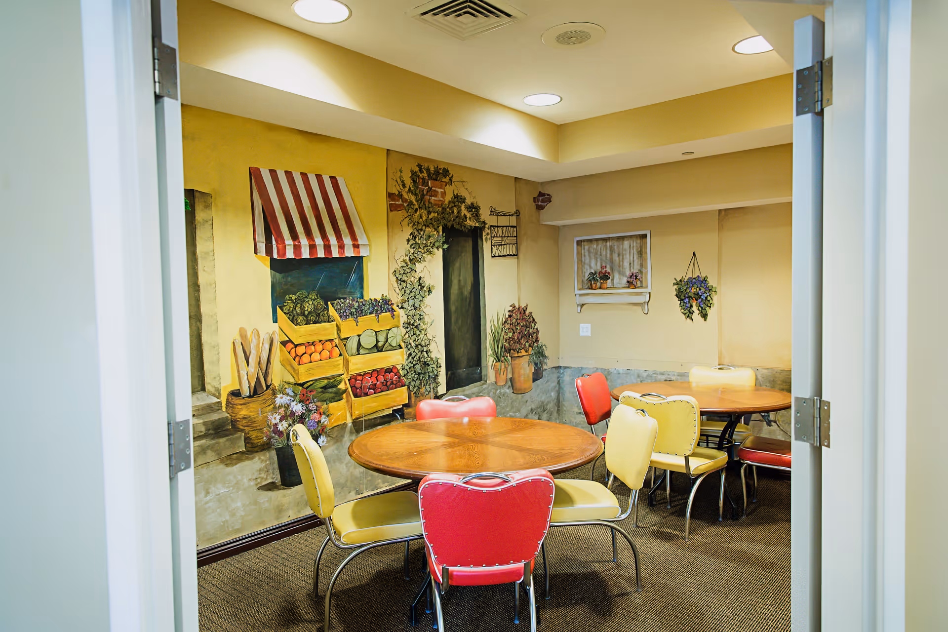 A small dining area with two round wooden tables surrounded by colorful chairs in red and yellow. The walls are decorated with a mural depicting a street market scene with crates of fruits and vegetables, potted plants, and a window with flowers. The room is well-lit with ceiling lights and has a carpeted floor.