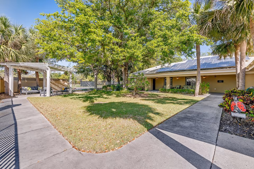 Outdoor courtyard area at Healthpark Senior Living featuring a grassy lawn with trees, a concrete walkway, a white pergola with seating underneath, and a building with solar panels on the roof in the background.