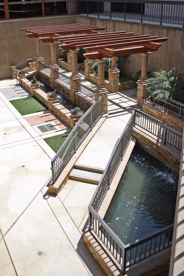 Outdoor courtyard area with a water feature including a narrow pond and small fountains, stone pillars supporting wooden pergolas, potted plants, and a concrete walkway with railings.