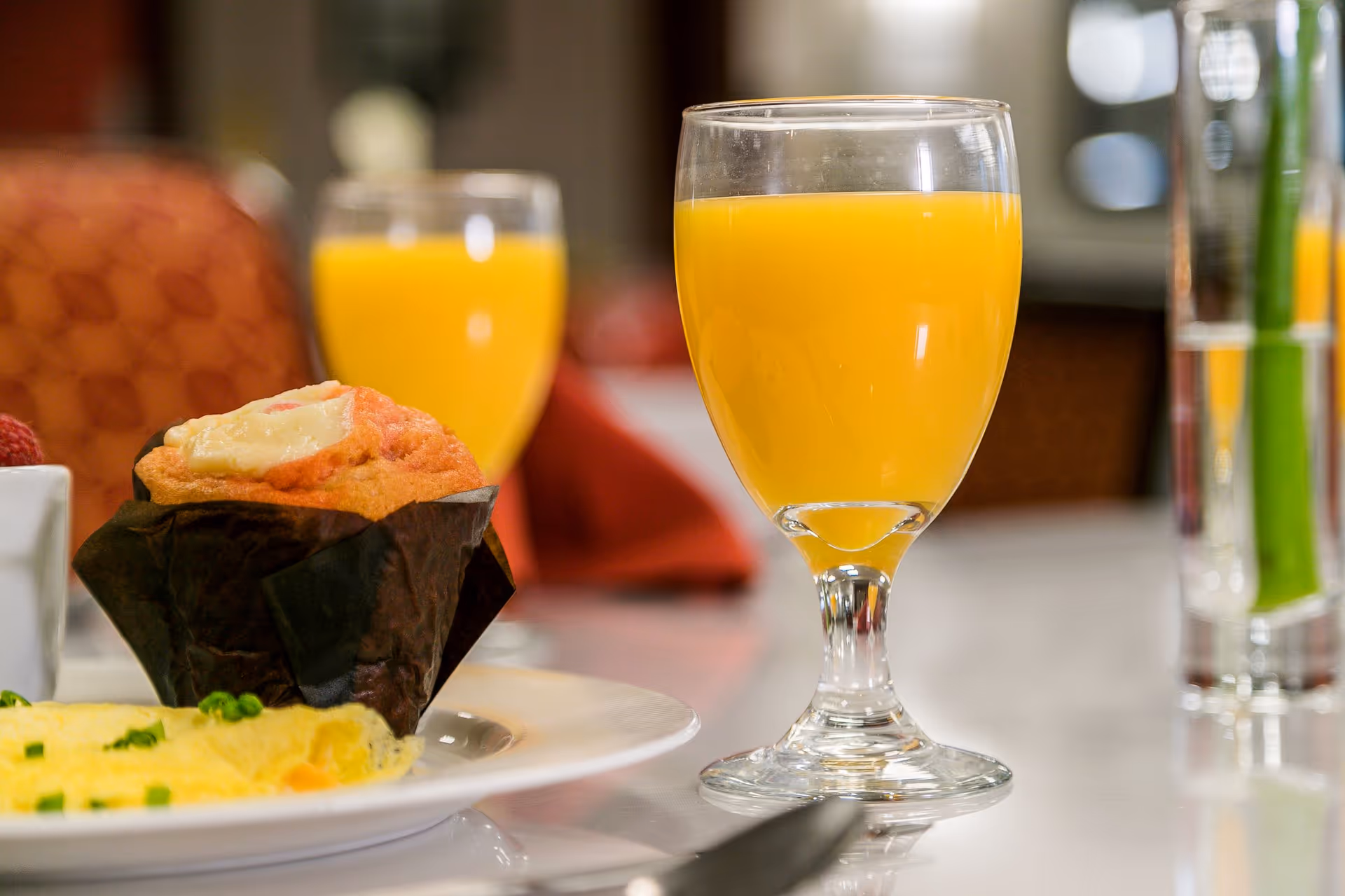 Close-up of a breakfast table setting with a glass of orange juice, a muffin in a dark wrapper, and a plate with an omelette garnished with chopped green onions. The background shows a blurred second glass of orange juice and a red chair.
