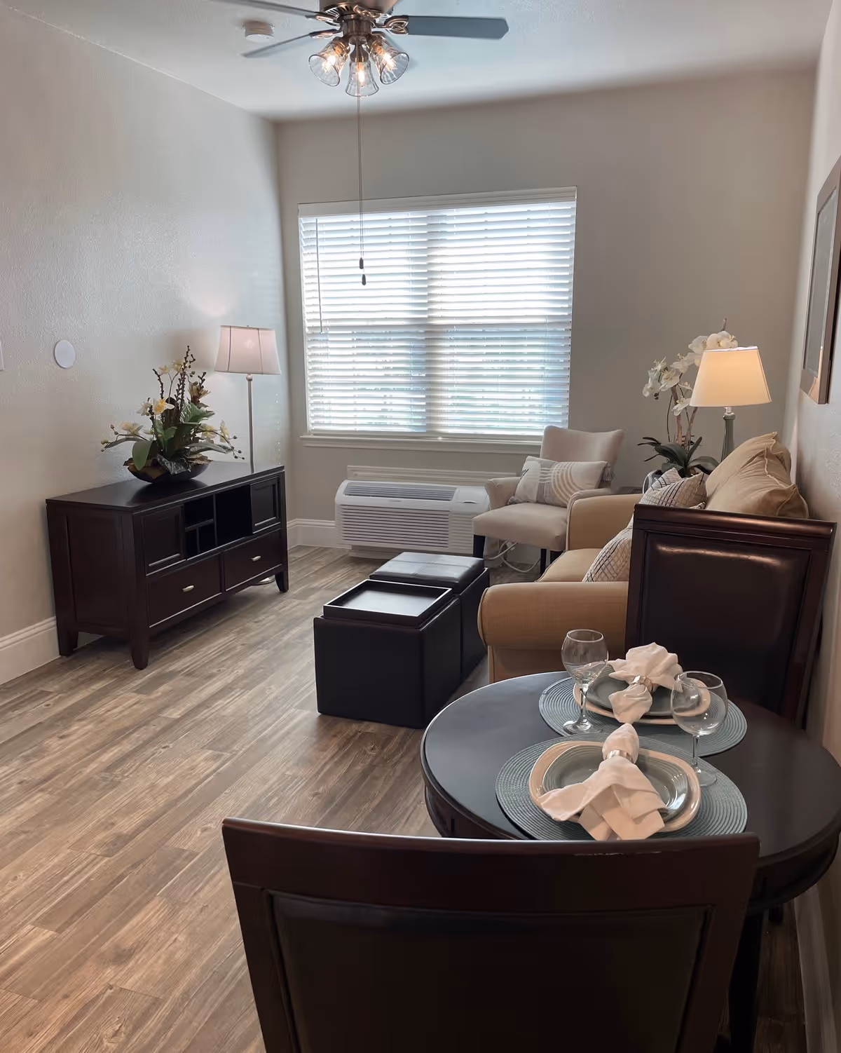 A cozy living room area with a beige sofa, an armchair, a dark wooden TV stand with a floral arrangement, a small round dining table set for two with plates, napkins, and wine glasses, and a window with blinds letting in natural light. The room has wood flooring and a ceiling fan with lights.