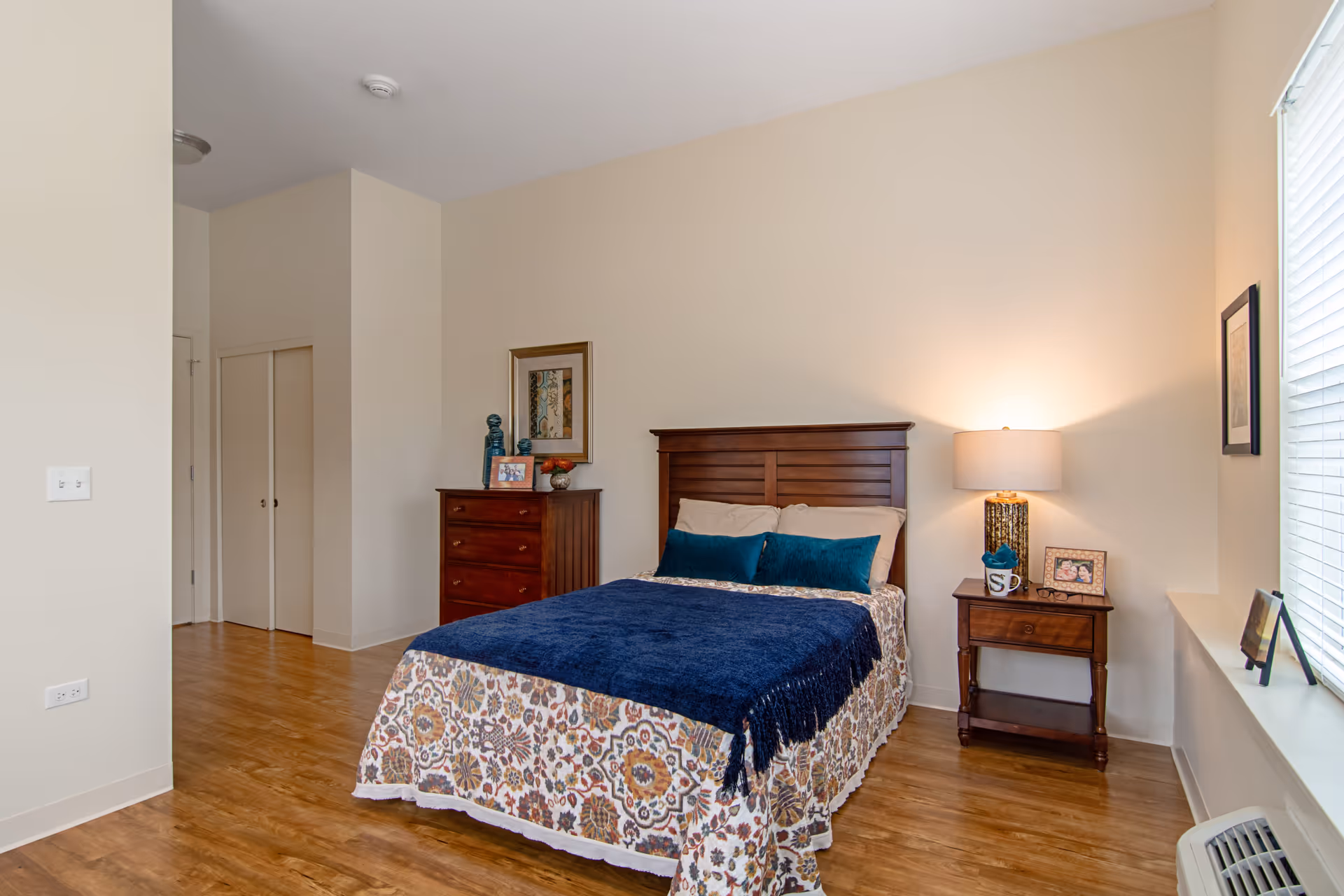 A bedroom with a wooden bed frame, a bed with a floral patterned bedspread and a dark blue blanket, two blue pillows, a wooden dresser with decorative items and a framed picture above it, a wooden nightstand with a lamp, a mug, and a framed photo, and a window with blinds letting in natural light.