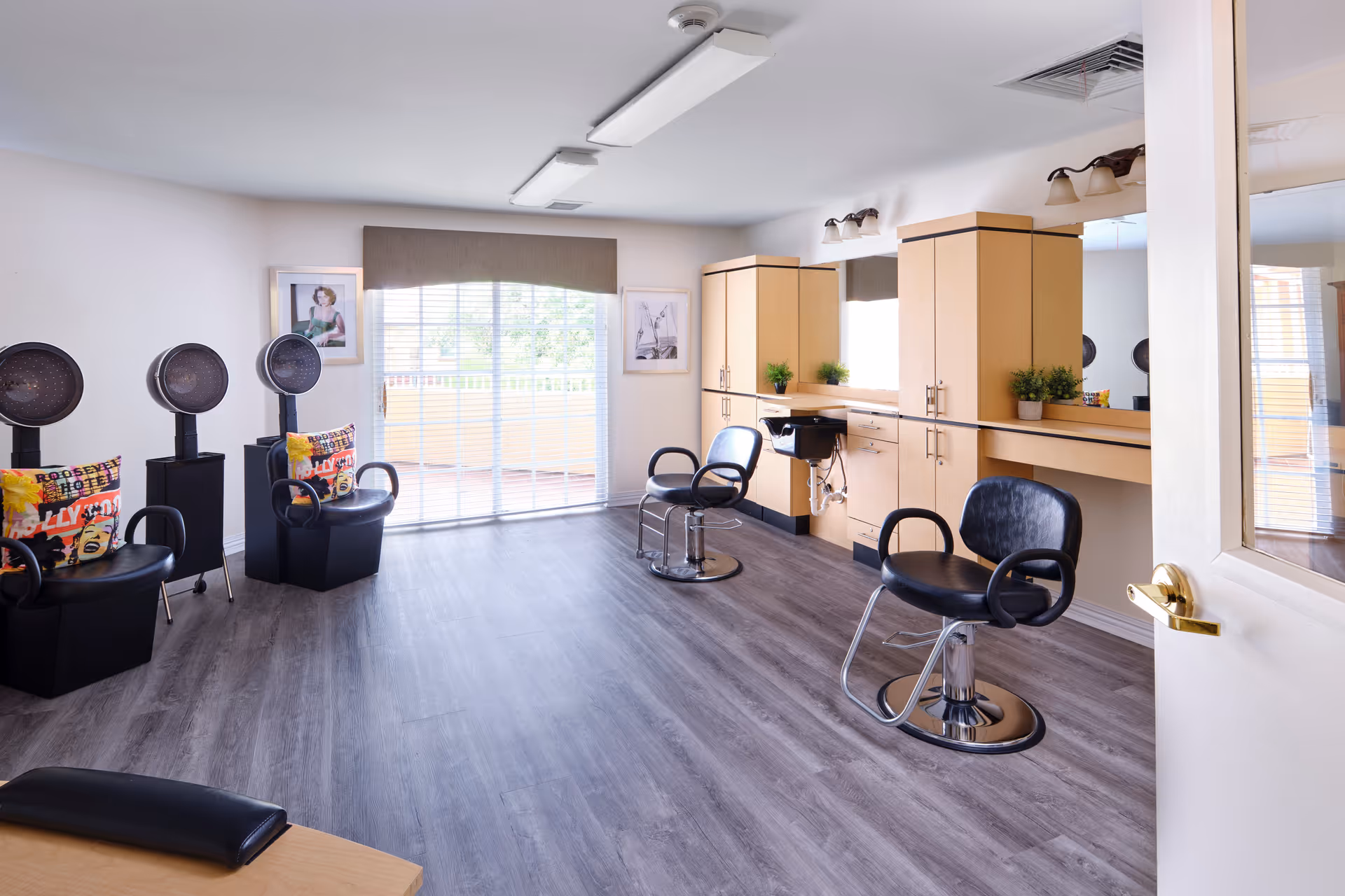 A bright and clean hair salon room with two black salon chairs in front of a large mirror and wooden cabinets. On the left side, there are three black hair dryers with colorful pillows on the chairs. The room has large windows with blinds letting in natural light and a door partially open on the right side.
