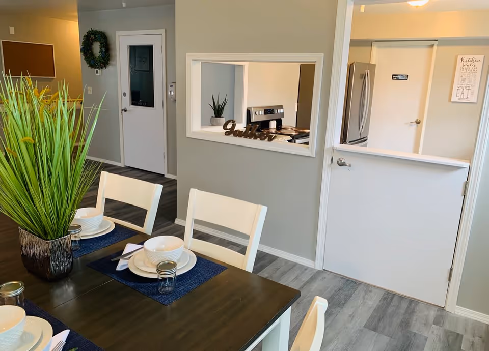 Dining area with a dark wooden table set with white bowls, plates, and small jars on blue placemats. White chairs surround the table. A decorative green plant is placed on the table. In the background, there is a kitchen visible through a pass-through window with a stainless steel stove and refrigerator. The walls are painted light gray, and there is a door with a wreath and a corkboard on the wall.