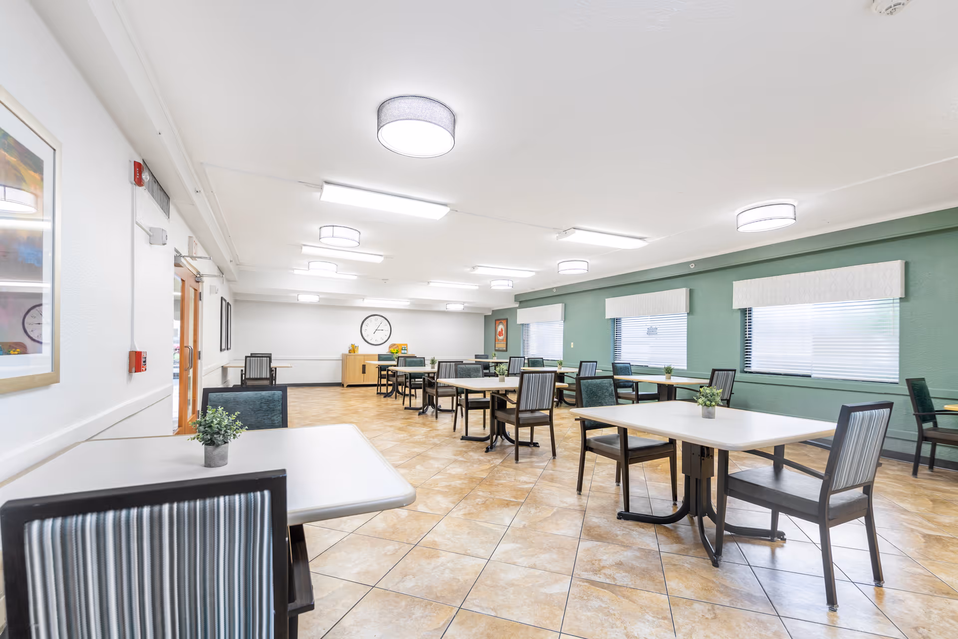 A spacious dining room with multiple tables and chairs arranged neatly. The room has tiled floors, green walls with windows covered by white blinds, and ceiling lights providing bright illumination. Small potted plants are placed on the tables, and a clock is visible on the far wall above a wooden cabinet.