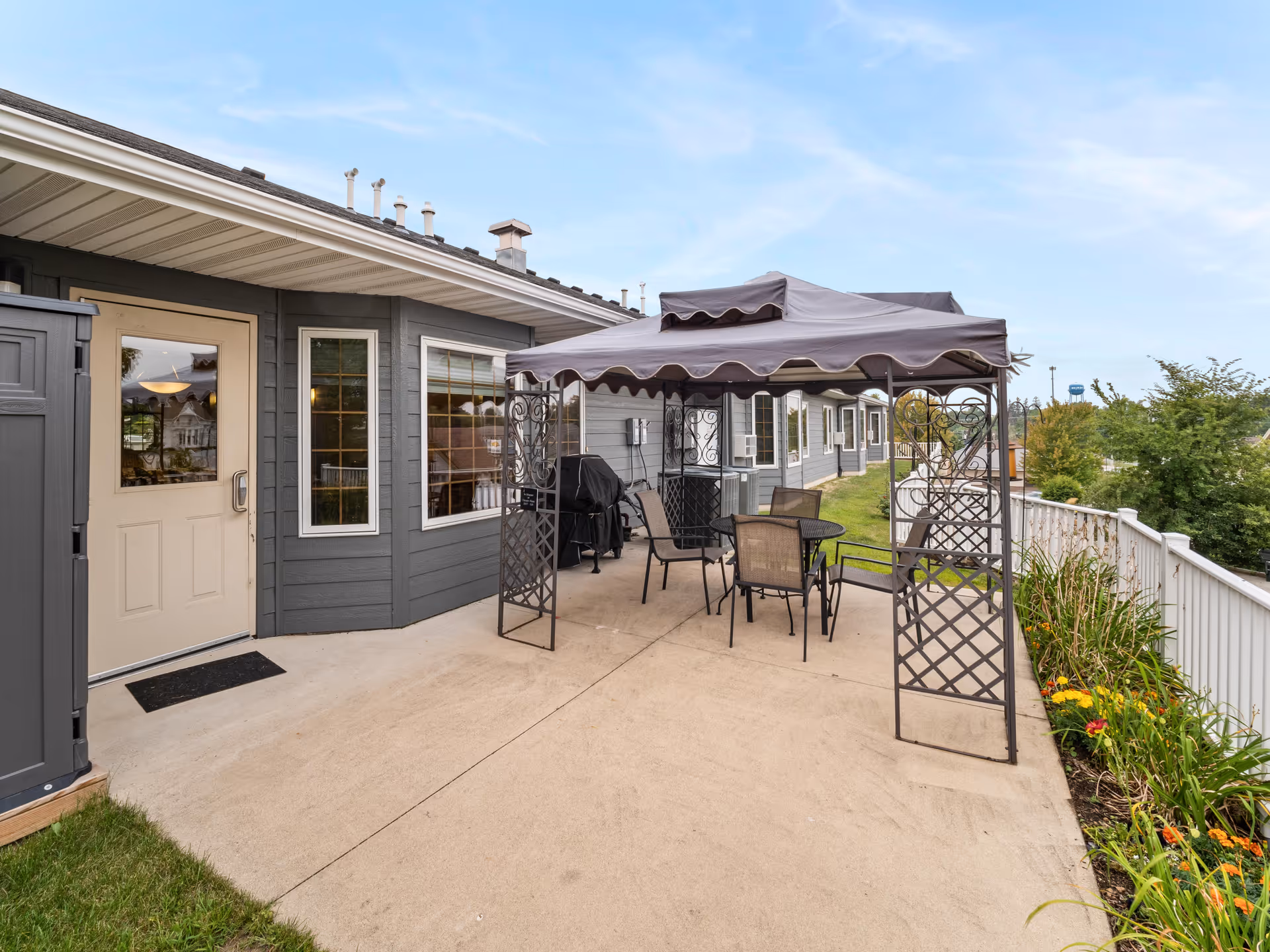Outdoor patio area at Good Samaritan Society - Waukon featuring a concrete floor, a metal gazebo with a fabric canopy, a round table with four chairs, a barbecue grill, and a garden bed with flowers along a white fence. The building exterior is gray with white trim and multiple windows.