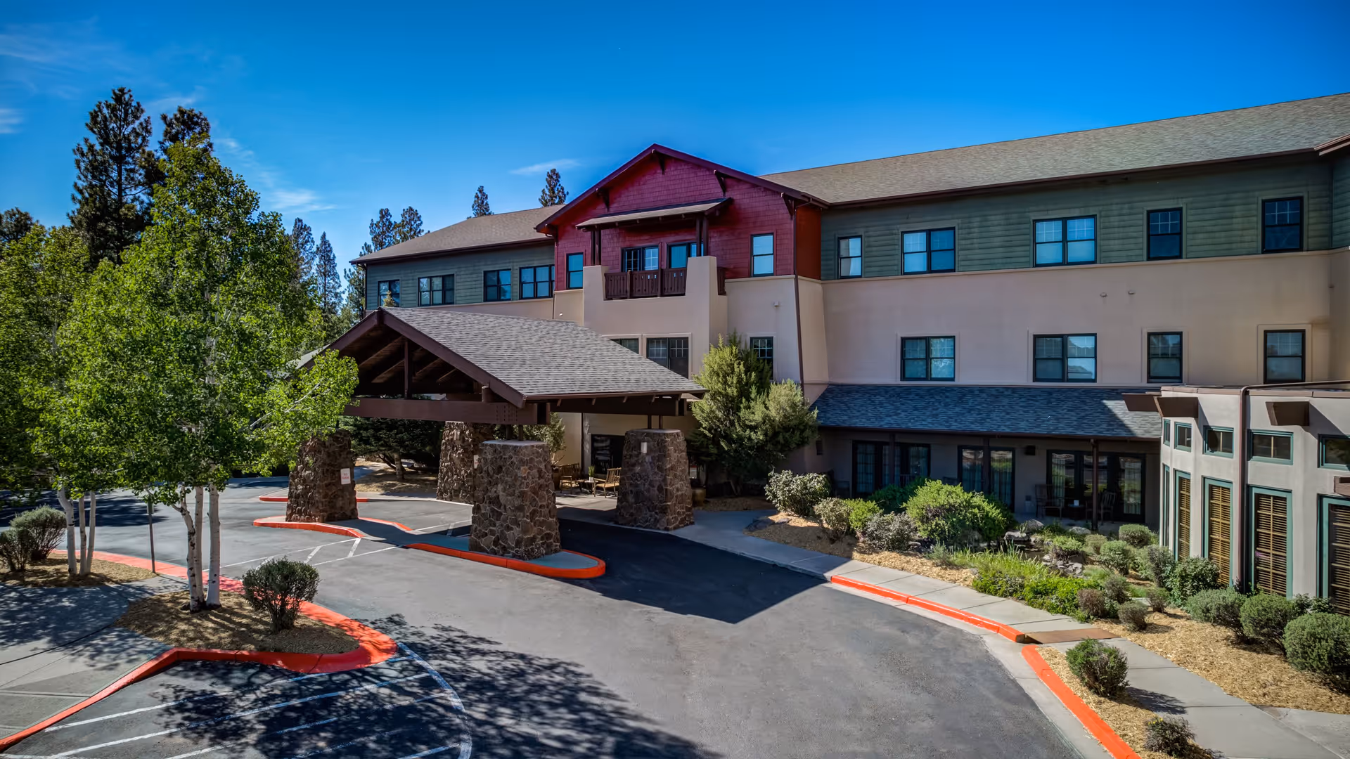 Exterior view of The Peaks, A Senior Living Community building with a covered entrance supported by stone pillars, surrounded by trees and landscaped bushes under a clear blue sky.