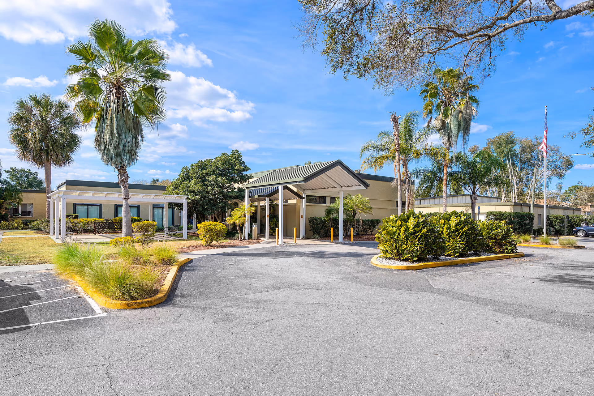 Exterior view of Ruleme Center showing a single-story building with a covered entrance, surrounded by palm trees, bushes, and a parking area under a partly cloudy blue sky.