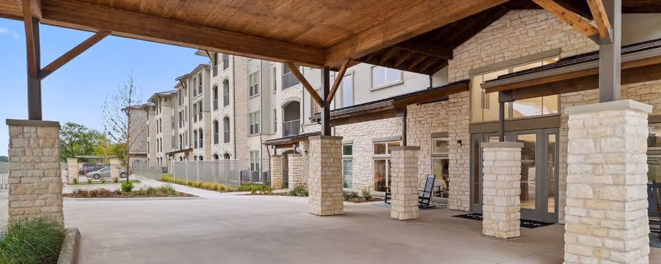 Covered stone-pillared porte-cochere and entrance to a multi-story senior living building with rocking chairs on the patio.