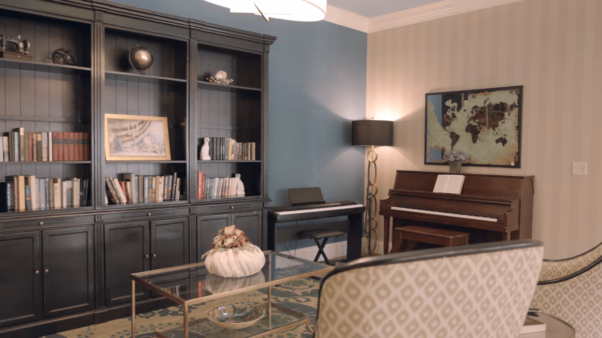 A cozy living room area featuring a large black bookshelf filled with books and decorative items, a glass coffee table with a white decorative centerpiece, a patterned armchair, an upright piano with sheet music, an electronic keyboard, a floor lamp, and a framed world map on the wall.