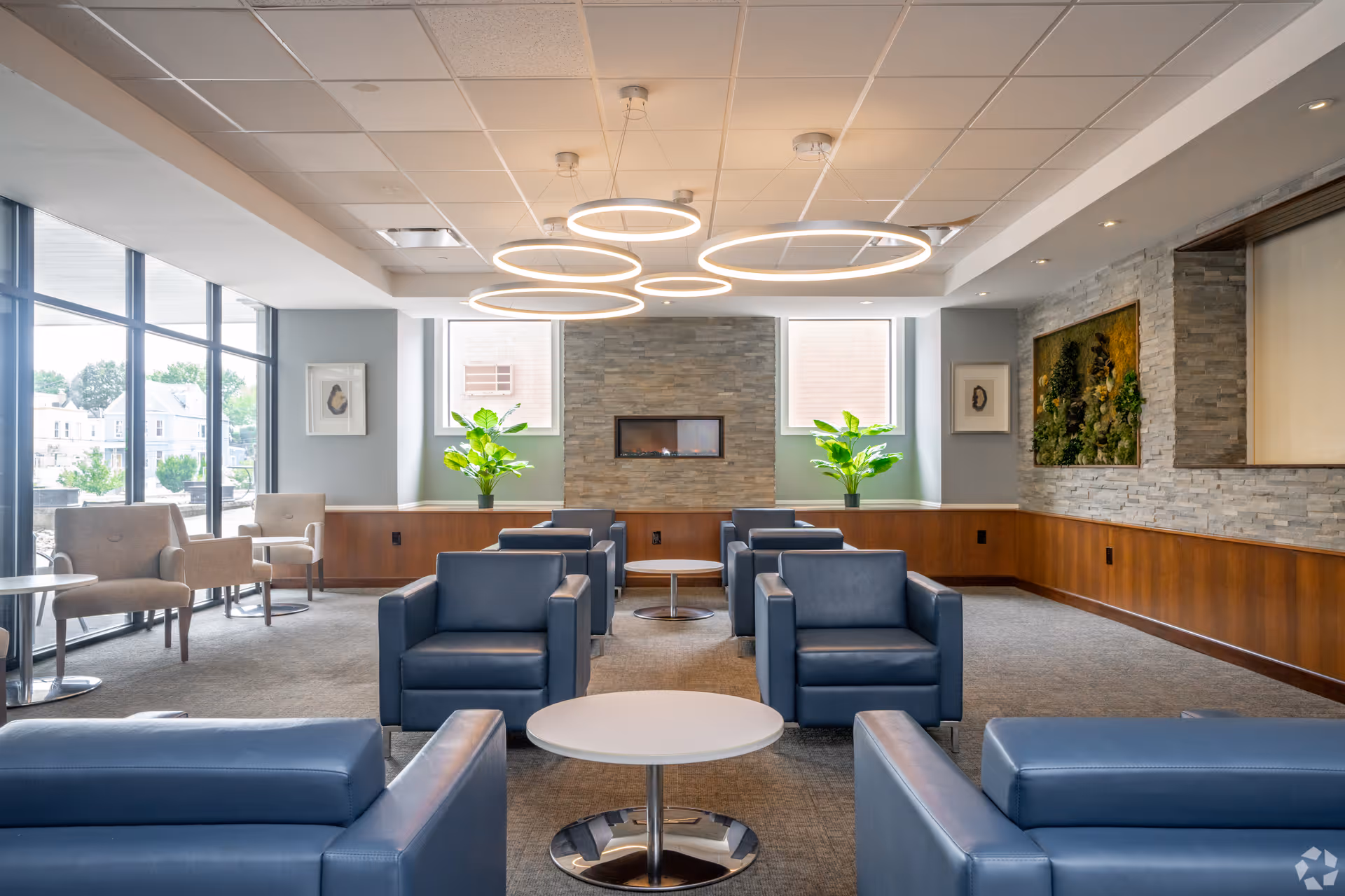 A modern senior living community lounge with blue armchairs and small round white tables arranged in the center. The room features large windows on the left side allowing natural light, a stone accent wall with a built-in fireplace, two green plants on a wooden ledge, and contemporary circular ceiling lights.