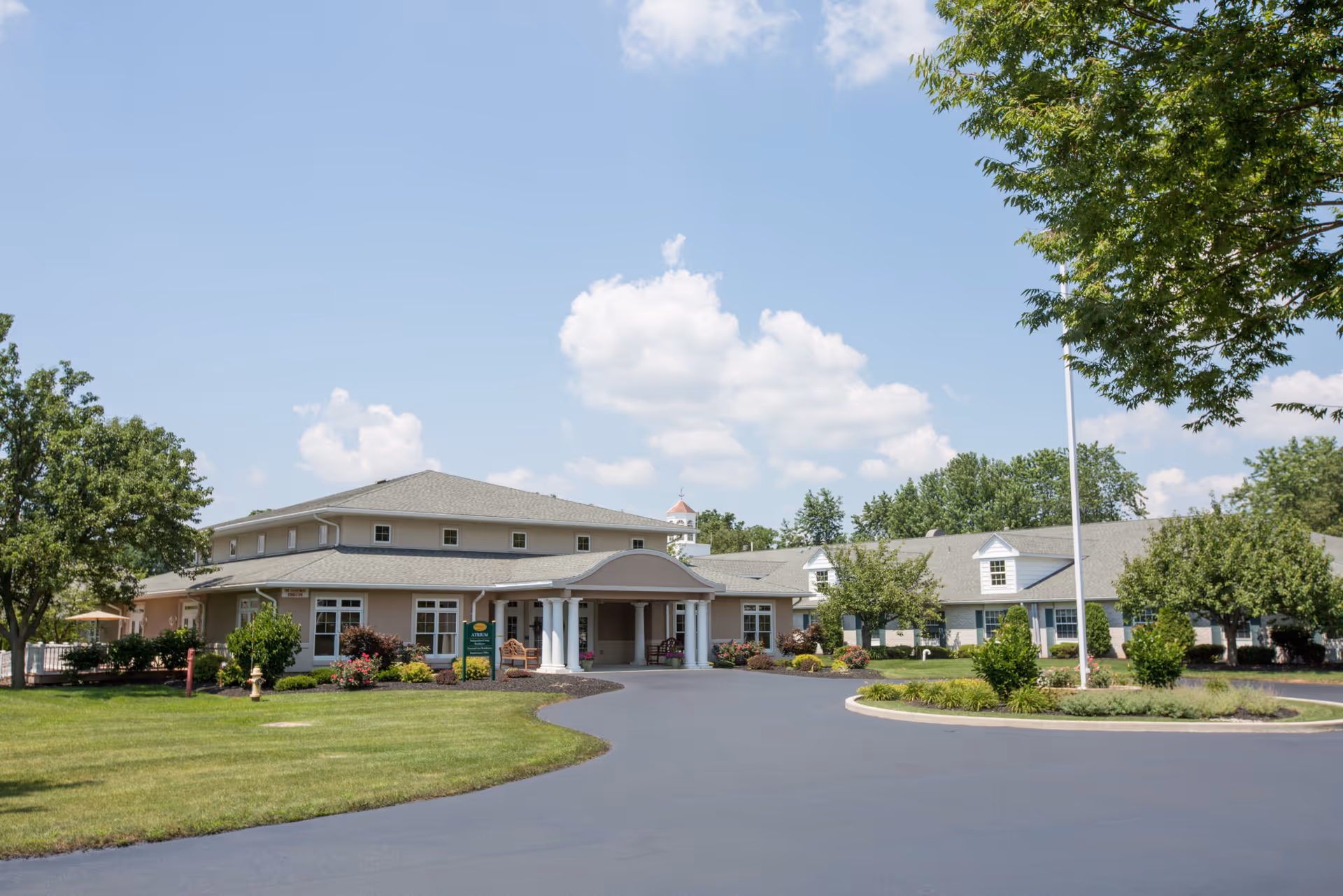 Exterior view of Country Meadows Of Bethlehem III, a single-story senior living facility with beige walls, white columns at the entrance, surrounded by green lawns, trees, and shrubs under a blue sky with scattered clouds.