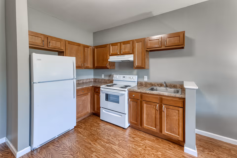 A kitchen with wooden cabinets, a white refrigerator, a white electric stove with oven, a range hood, a stainless steel sink, and brown countertops. The floor is wood-patterned, and the walls are painted light gray.