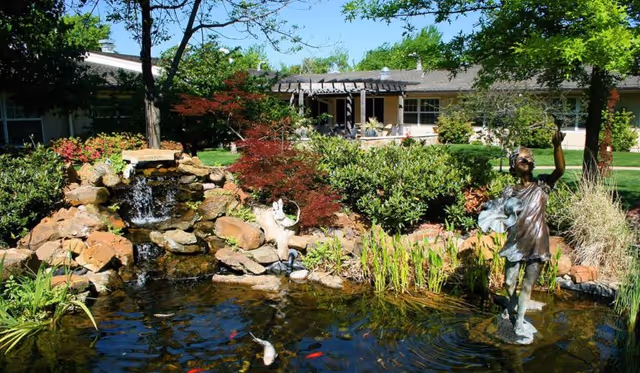 A serene outdoor garden area at Saint Simeon's Senior Community featuring a small waterfall cascading over rocks into a pond with koi fish. The pond is surrounded by lush greenery, including bushes, trees, and ornamental plants. A bronze statue of a child holding a bird stands in the pond, and a building with a pergola is visible in the background under a clear blue sky.