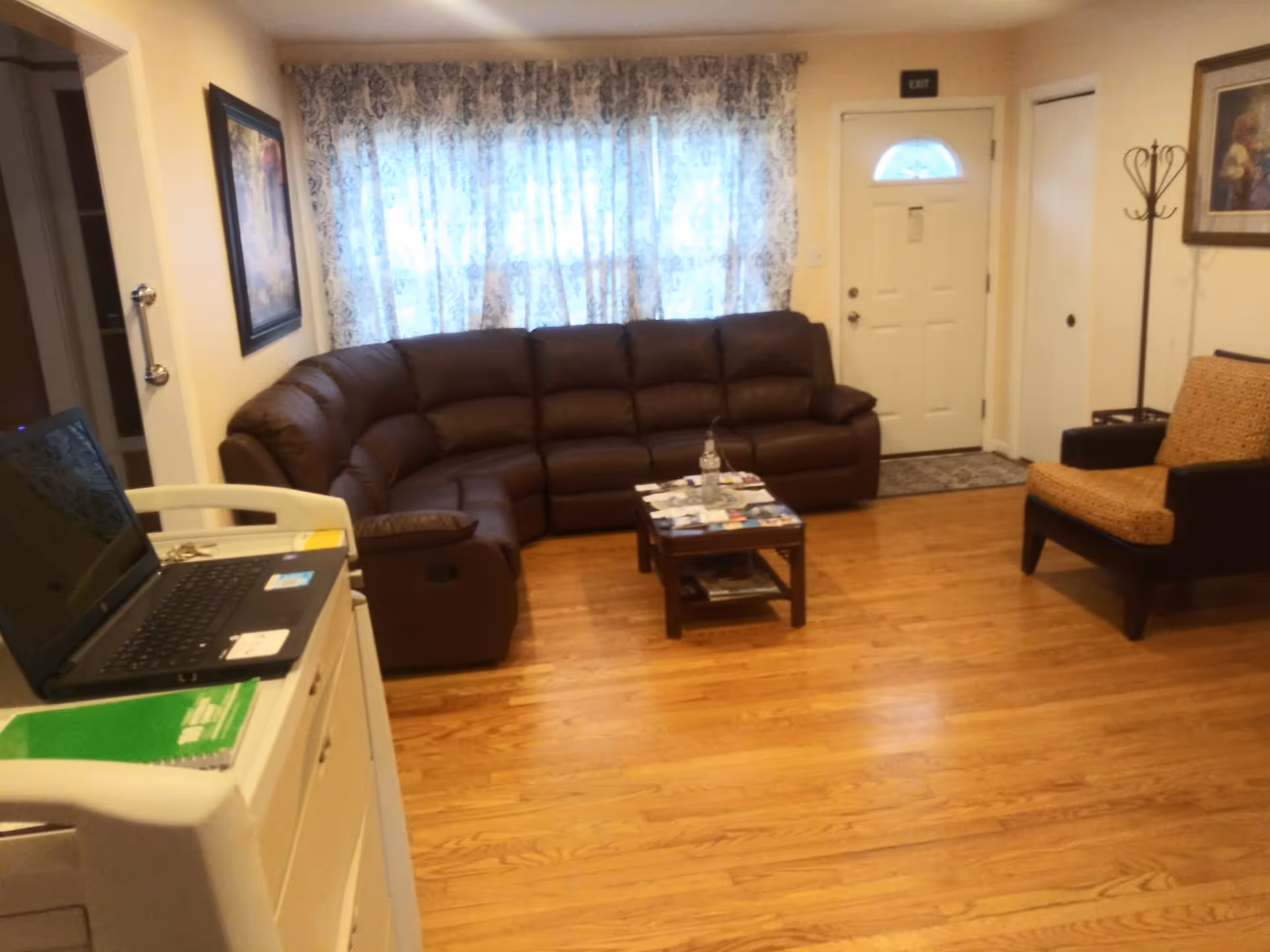 A cozy living room with a large brown sectional sofa, a small wooden coffee table with magazines and a water bottle on it, a single armchair with patterned upholstery, hardwood floors, a window with patterned curtains, a white door with an exit sign above it, and a laptop on a white cart in the foreground.