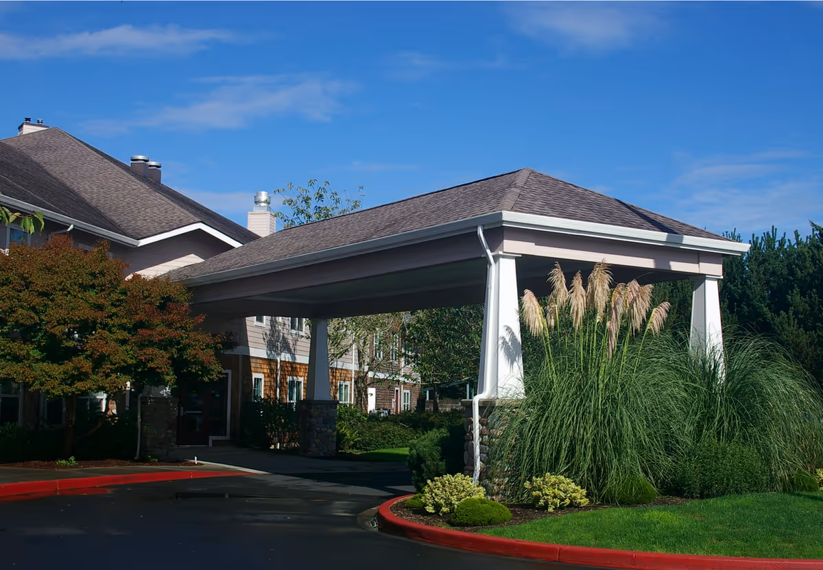 Covered entrance area of a senior living facility with a pitched roof supported by white columns, surrounded by well-maintained landscaping including tall grasses and bushes, under a clear blue sky.
