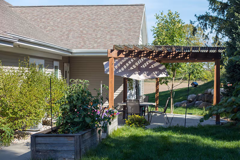 Outdoor patio area with a wooden pergola, umbrella-shaded table and chairs, raised garden beds, and lawn beside the building.