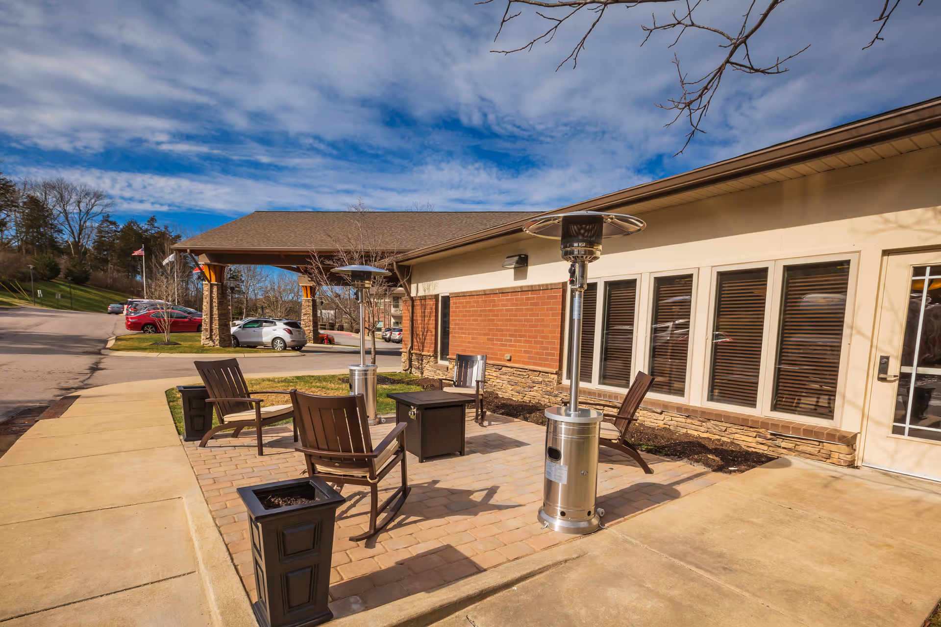 Outdoor patio area at The Lodge at Natchez Trace with several brown wooden chairs arranged around a small table, two tall outdoor heaters, and a brick-paved seating area next to a building with large windows and a door. Cars and trees are visible in the background under a partly cloudy sky.