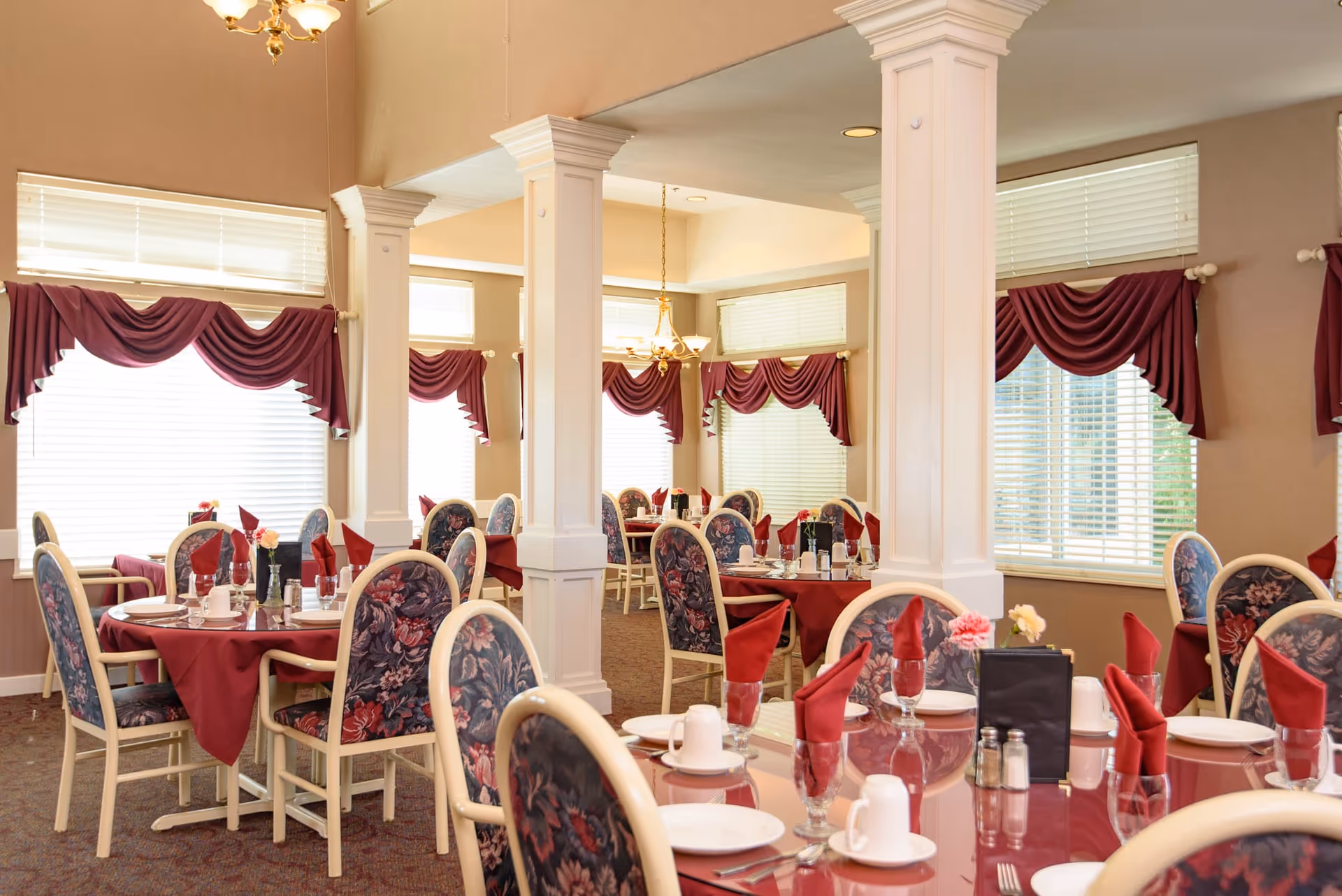 A bright dining room with multiple round tables set with red napkins, floral-upholstered chairs, white columns and large windows with burgundy swags.