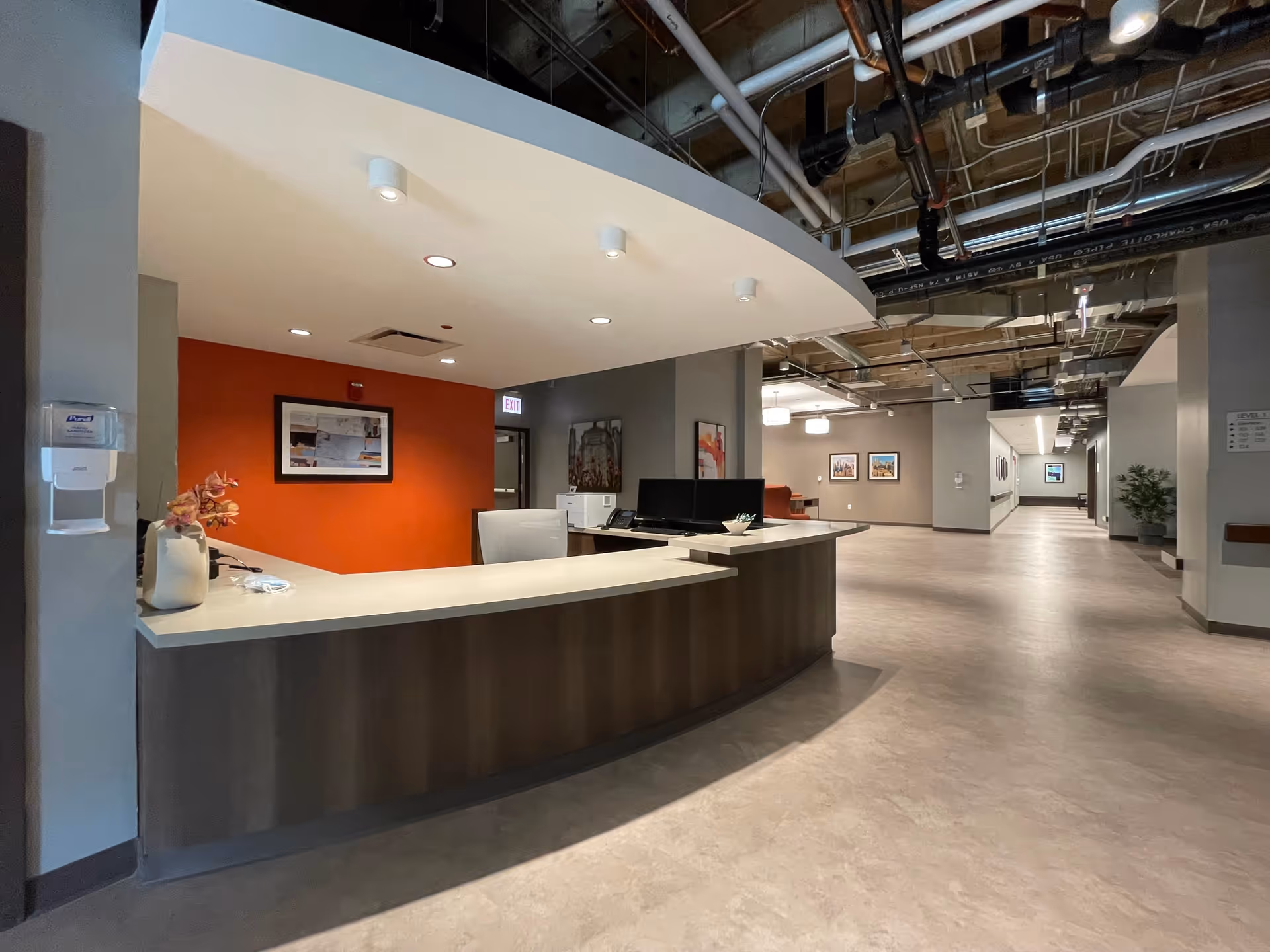 Reception desk area inside Ravenswood Supportive Living facility with a curved counter, a white chair, a vase with flowers, and computer monitors. The background features a red accent wall with framed artwork, an exit sign, and a wide hallway with additional framed pictures and plants.