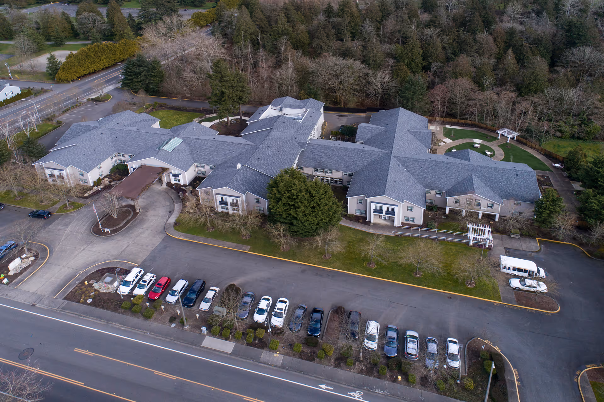 Aerial view of a large senior living facility building surrounded by trees and greenery. The building has multiple wings with gray roofs and beige walls. There is a parking lot with several cars parked in front of the building, and a circular driveway at the entrance. A small garden area with a white pergola is visible on the right side of the building.