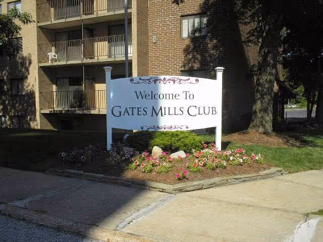 Outdoor view of the entrance sign for Gates Mills Club Senior Living, surrounded by flowers and greenery, with a multi-story brick building and balconies in the background.