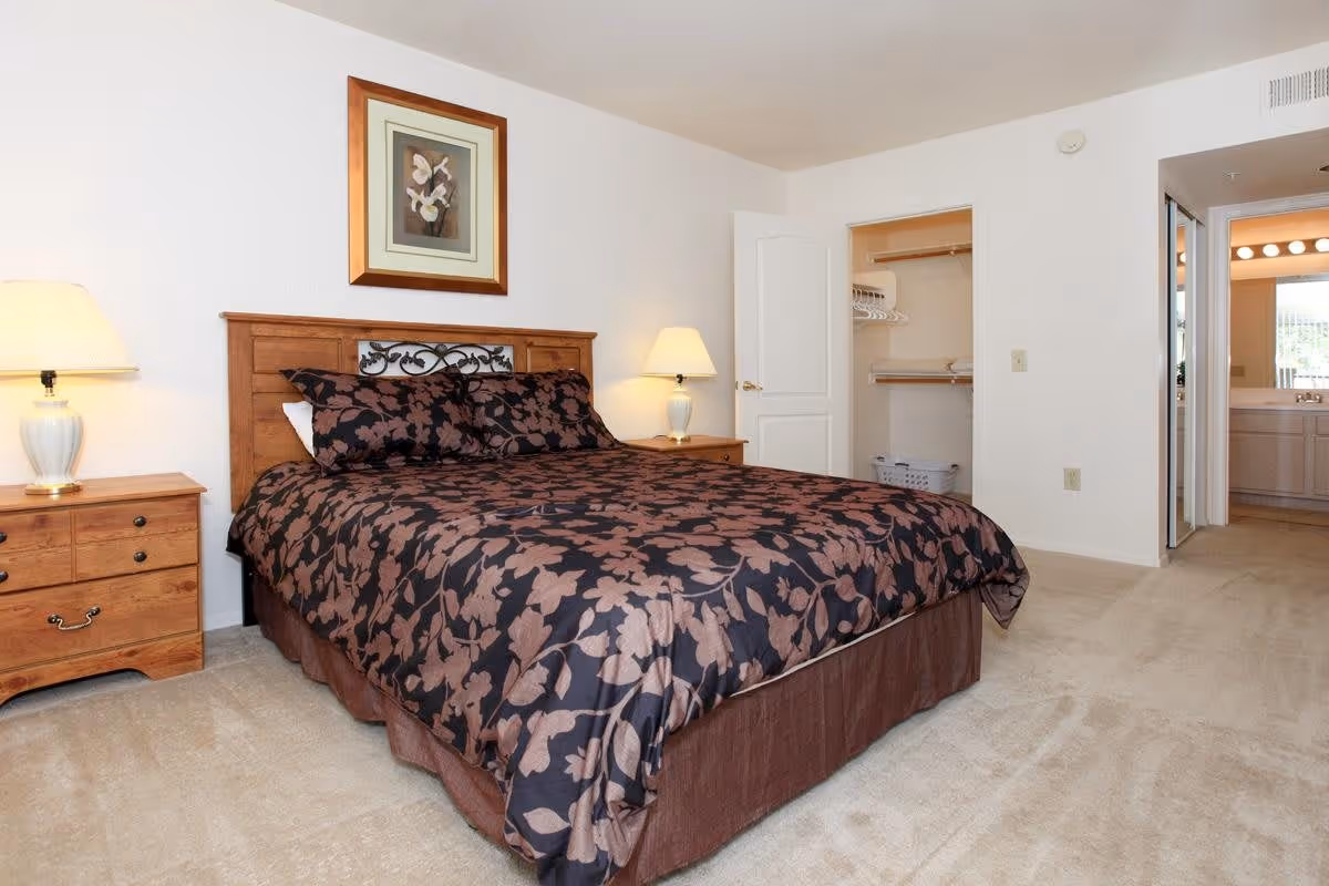 A bedroom with a large bed covered in a dark floral patterned comforter. There are two wooden nightstands on either side of the bed, each with a lamp. A framed floral picture hangs on the wall above the headboard. An open door reveals a closet with hangers and a laundry basket. Another open doorway leads to a bathroom with a vanity and mirror.