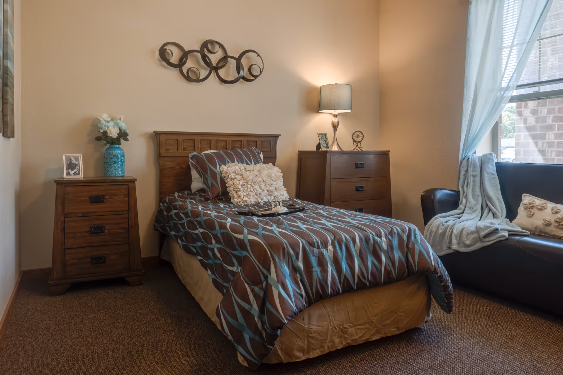 A cozy bedroom with a single bed covered in a brown and blue patterned comforter and decorative pillows. There is a wooden nightstand with a vase of flowers and a framed photo on the left side of the bed, and a wooden dresser with a lamp, a small clock, and a framed picture on the right side. A black leather couch with a light blue throw blanket and a decorative pillow is positioned near a window with sheer curtains, allowing natural light to enter the room. The walls are painted beige and decorated with a circular metal wall art piece above the bed.