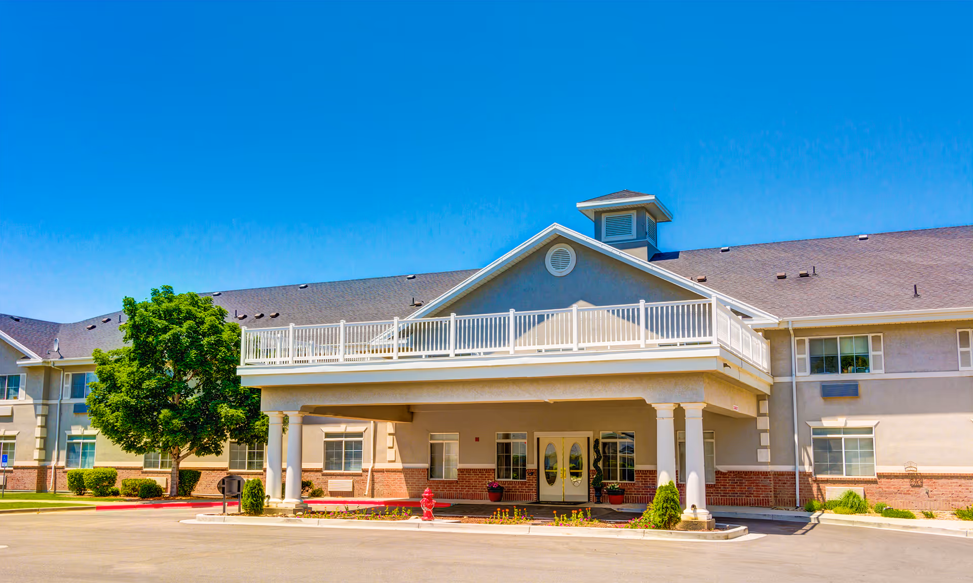 Exterior view of a two-story senior living facility building with a covered entrance supported by columns, a balcony above the entrance, several windows, a tree, and a clear blue sky.