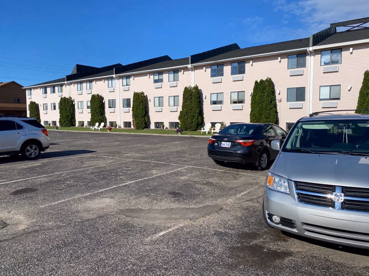 Exterior view of a three-story senior living facility building with light-colored siding, multiple windows, and tall evergreen shrubs in front. Several cars are parked in the asphalt parking lot in front of the building under a clear blue sky.